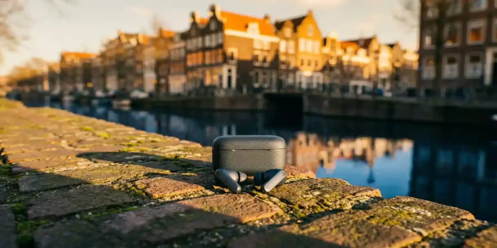 Draadloze oordopjes op een verweerde bakstenen richel langs een Amsterdamse gracht, met grachtenpanden in zachte gouden-uurlicht op de achtergrond.