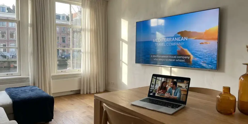 Flatscreen tv aan witte muur en laptop op eiken bureau tonen videoadvertenties, warm middaglicht via Nederlandse ramen met grachtzicht.